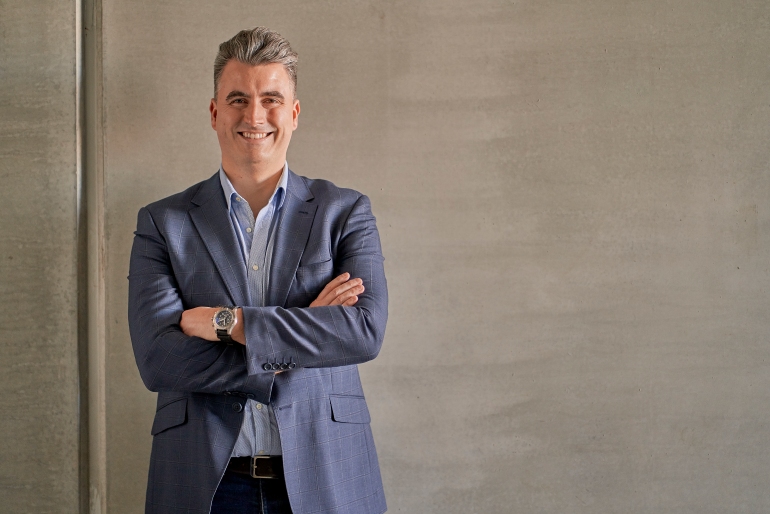 Matt is standing against a beige wall background, wearing a blue suit, with his arms folded. His blonde-streaked short hair is stylishly combed back and he is smiling.