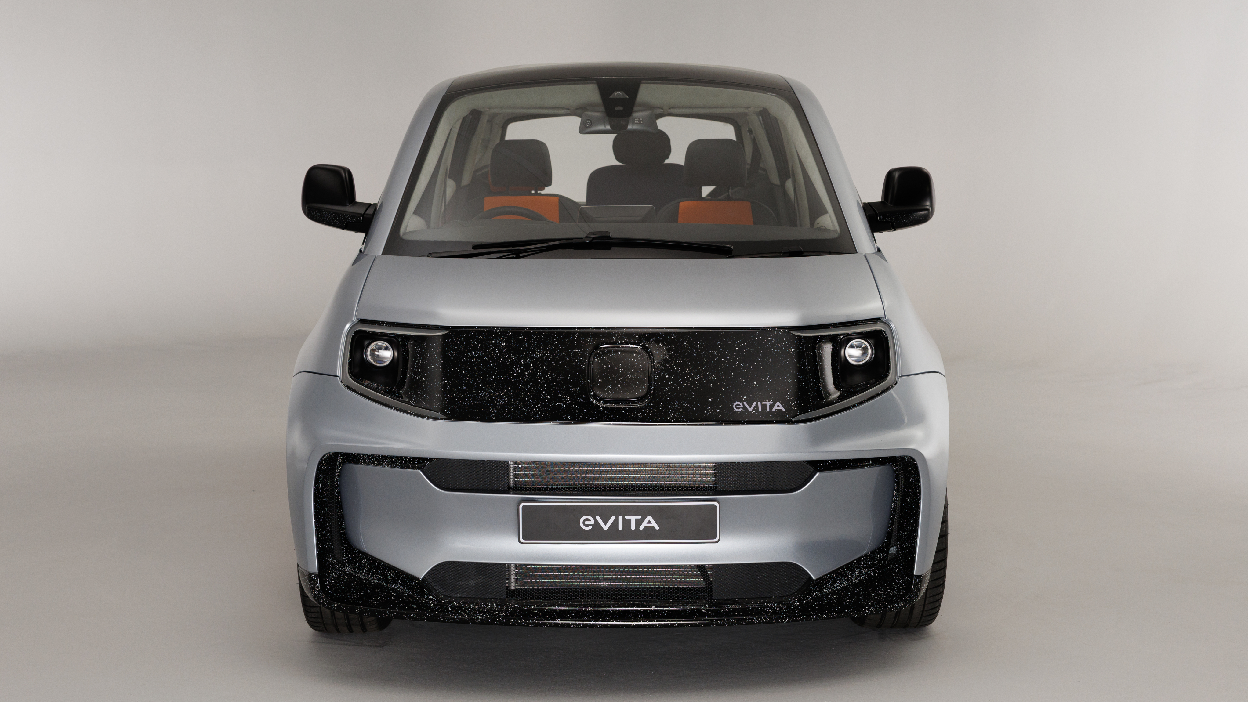 Front view of the e-VITA. The car features a smooth silver body with a black speckled front panel and integrated headlights, photographed in a studio setting.