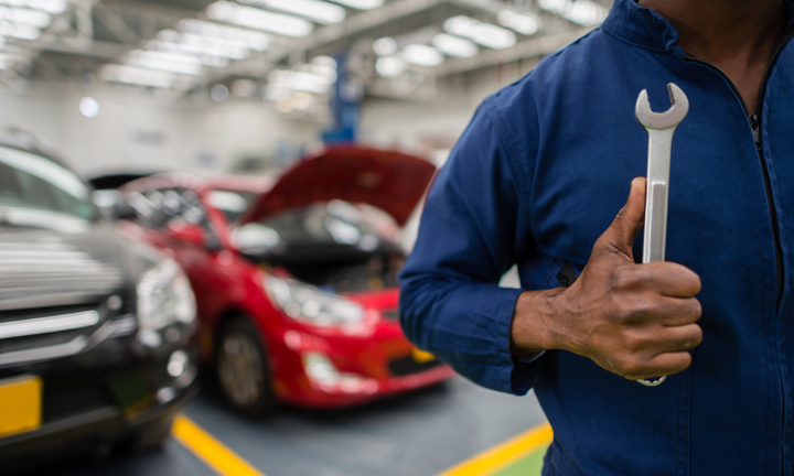 Technician in a blue coverall holding a wrench in an auto repair shop, with cars in the background.