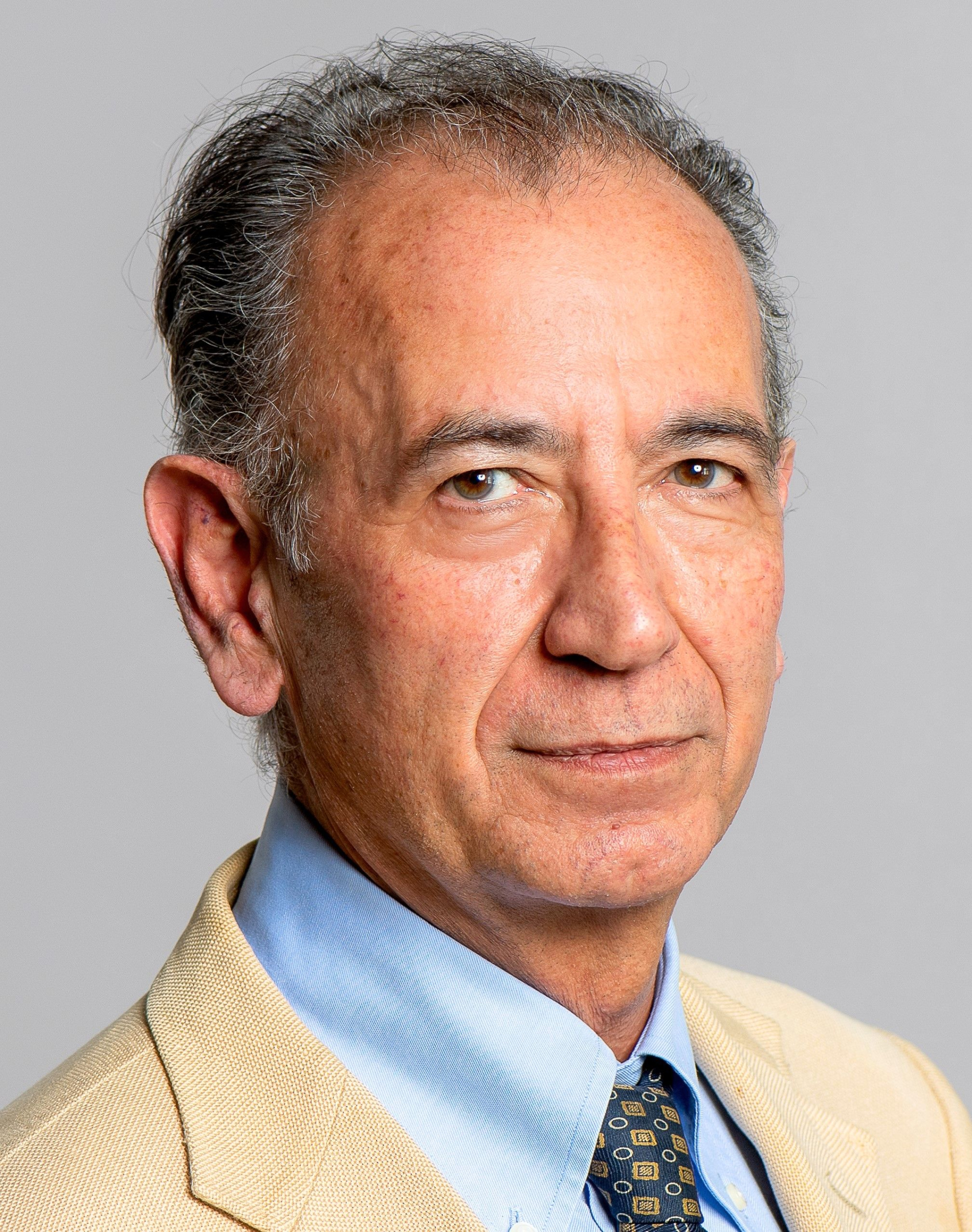 Portret mężczyzny na neutralnym, jasnym tle. Ma krótkie, siwiejące włosy i brązowe oczy, ubrany jest w jasną marynarkę, błękitną koszulę i wzorzysty krawat. Ujęcie do ramion, twarz zwrócona lekko w bok, spokojny, poważny wyraz.